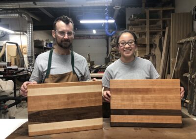 Two Participants in the woodshop showing off their handcrafted cutting boards in the woodshop, both dressed for safety