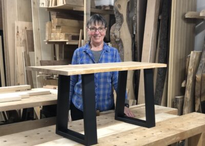Smiling LCW member proud of her new design for a bench, pale seat with black legs, in the woodshop surrounded by wood