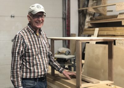 Smiling older LCW member proud of his wooden table in the woodshop with wood on the shelves