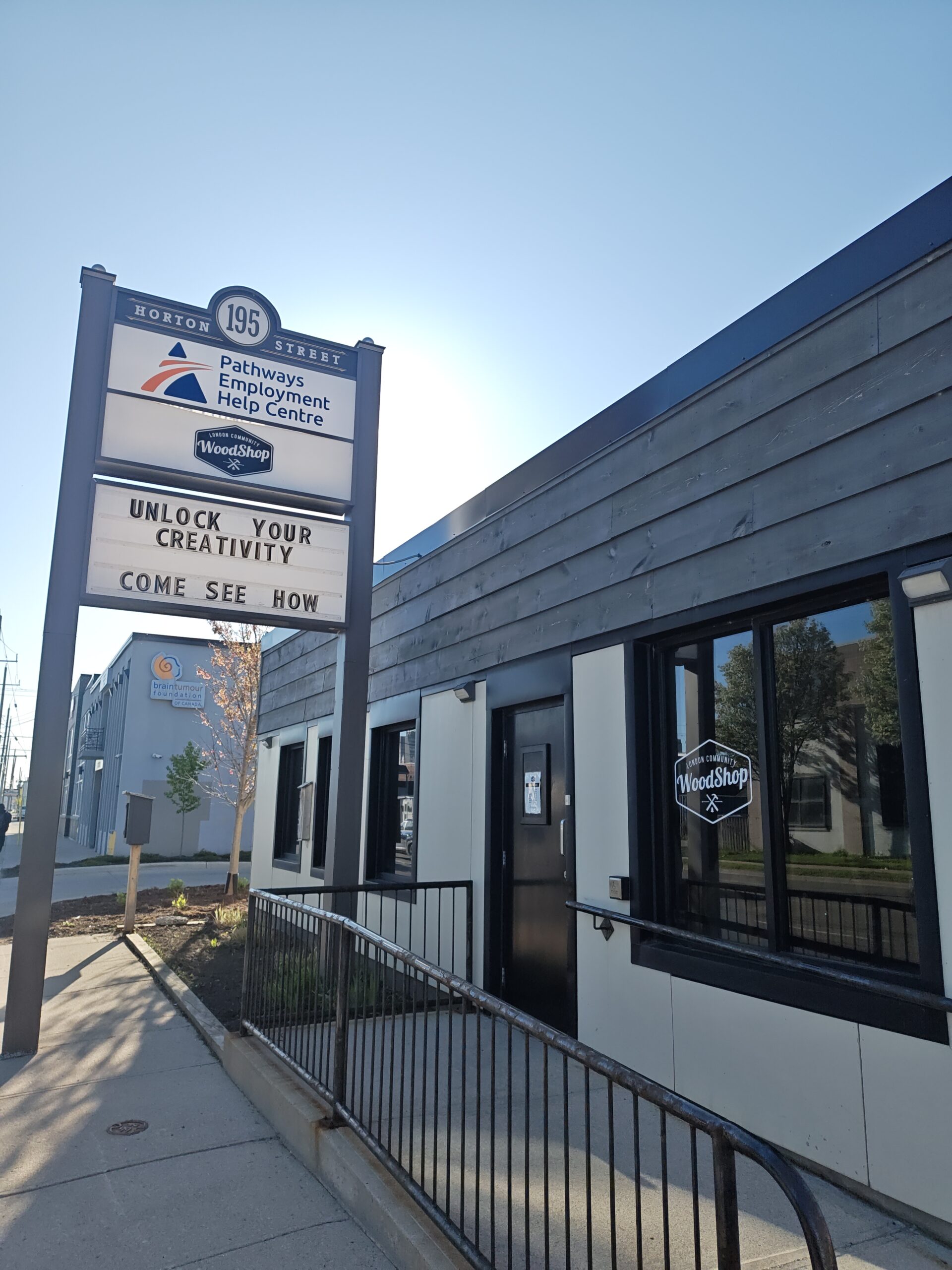 London Community Woodshop outdoor sign next to the woodshop building. 2 logos, Pathways Employment Help Centre and London Community Woodshop above text sign that says 'unlock your creativity. Come see how'